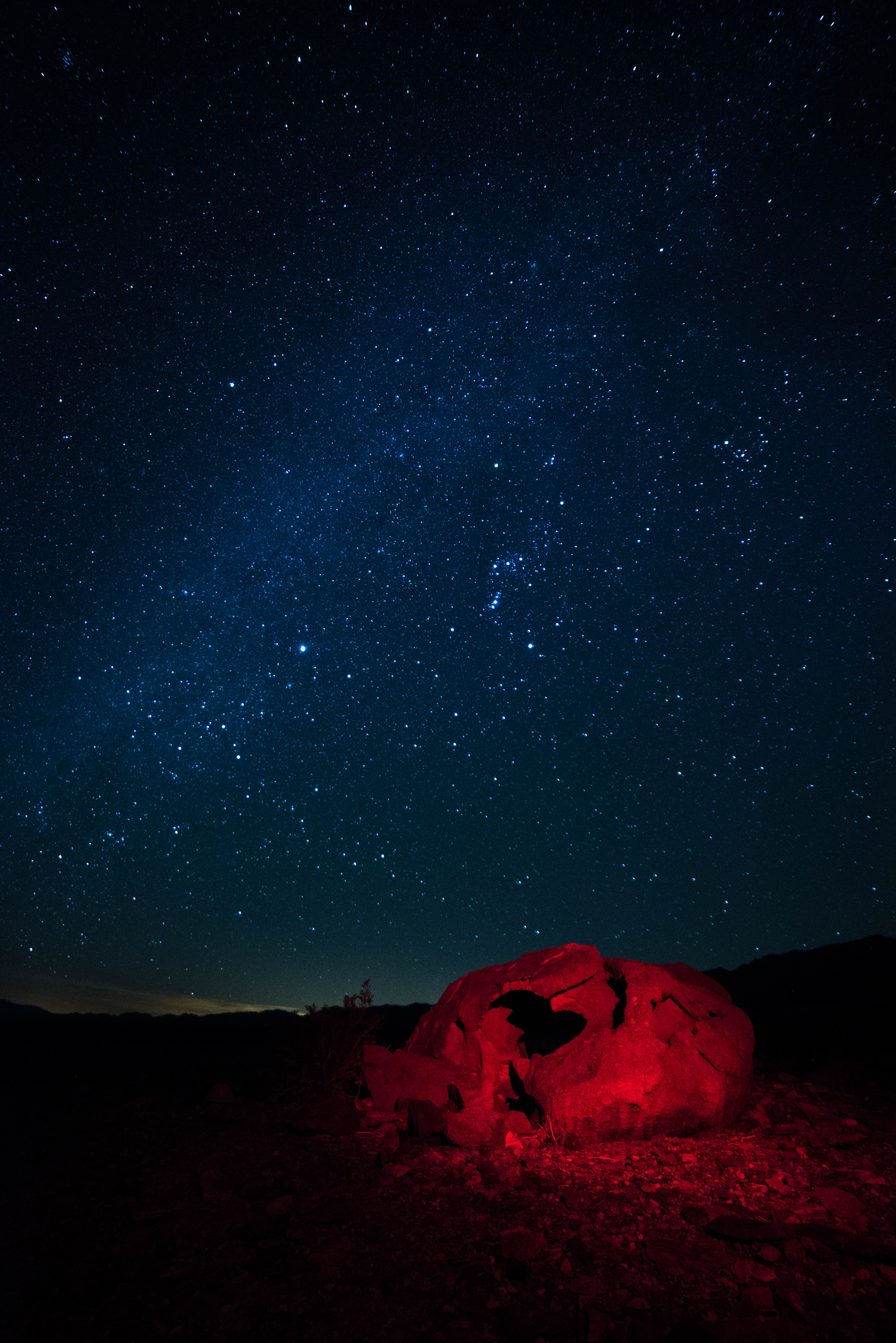 The night sky shines brightly behind a red rock that is lit with a red light. In the distant horizon, the hazy orange light of Las Vegas is visible.