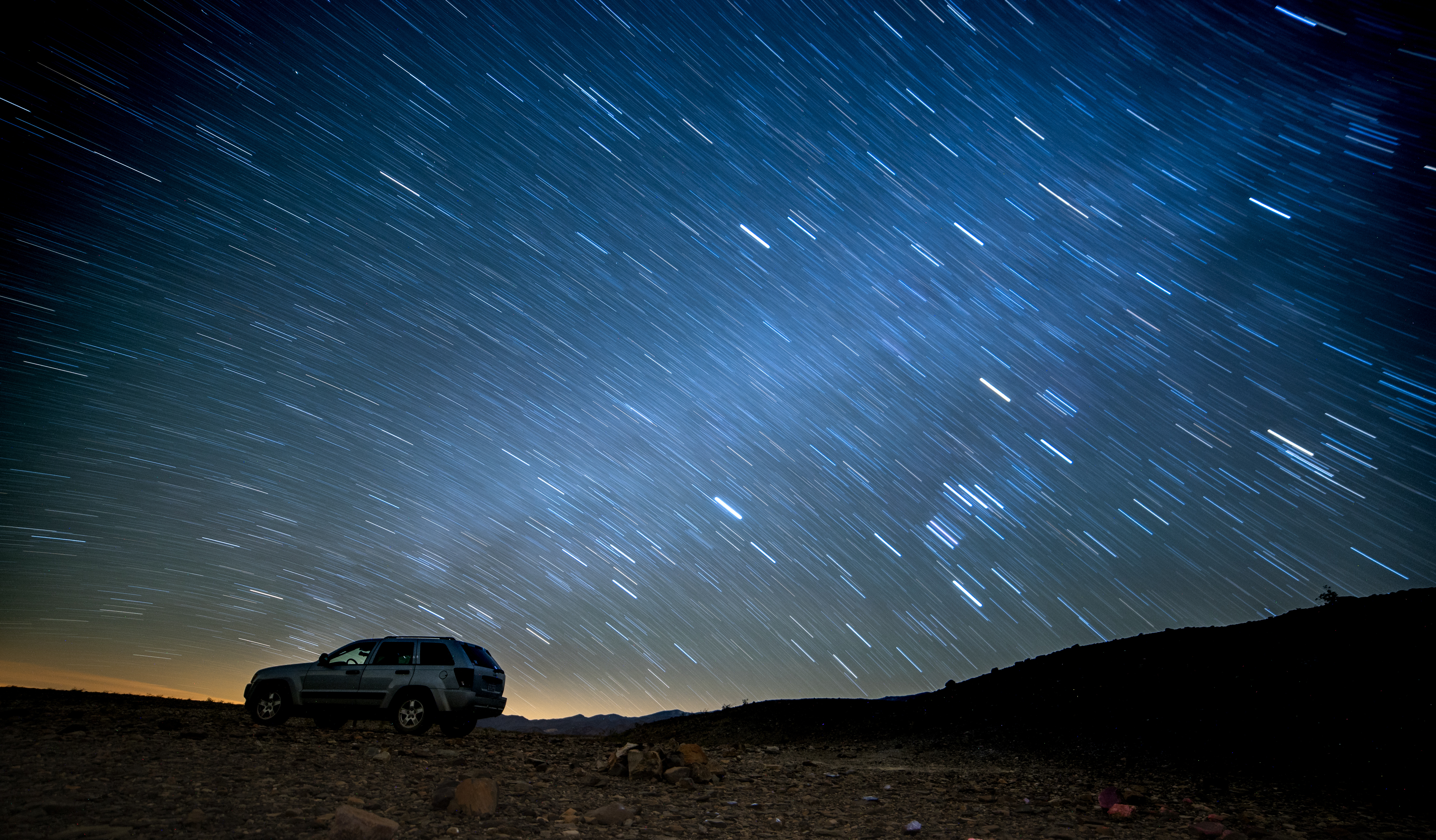 A bright lit night sky with star trails - streaks of white and grey. In the foreground a Jeep is parked on a campsite.