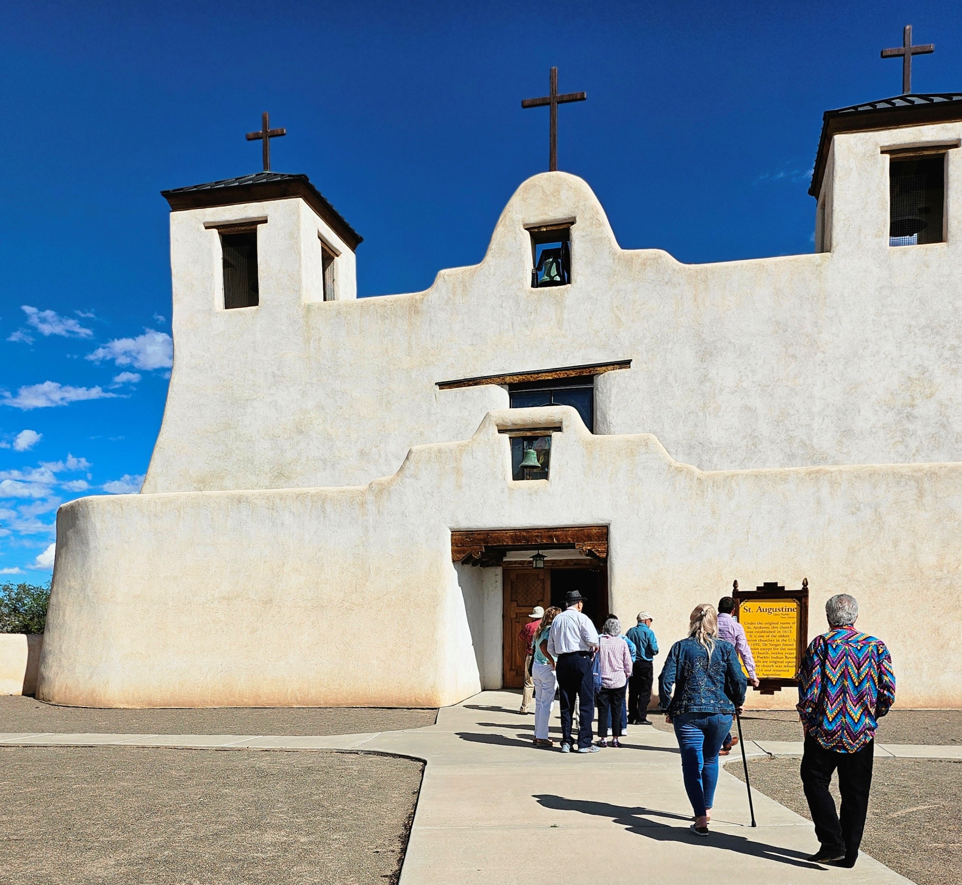 A group of people walk toward the entrance of a large cream-colored adobe church. There is a short wall in front with a bell hanging in the center and a larger wall behind with a second bell hanging from an archway and three crucifixes.