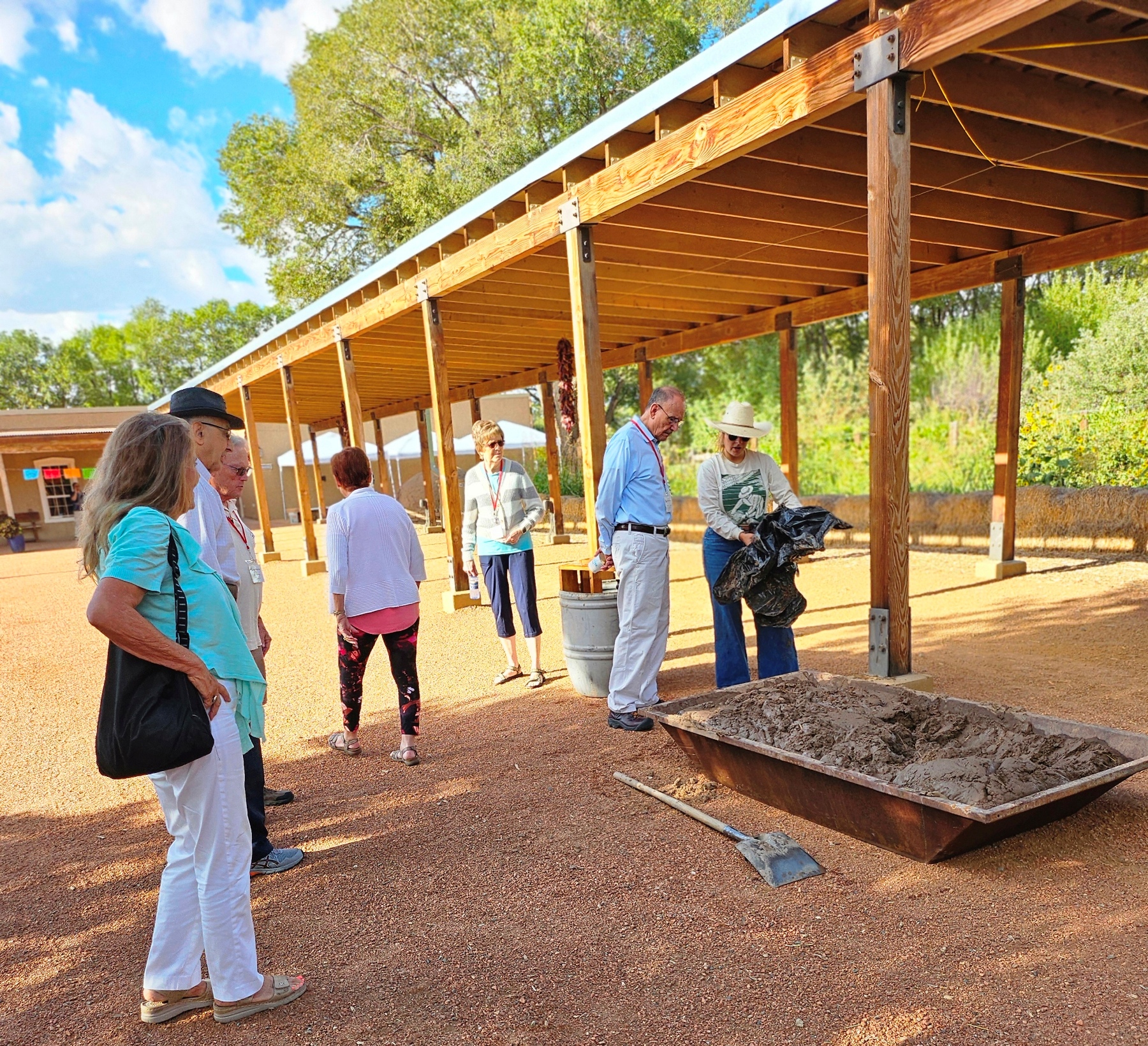 A group of people gather outside on the red gravel near a long shade structure while a woman with a cowboy hat holds a black plastic bag over a rectangular trough filled with mud with a shovel next to it.