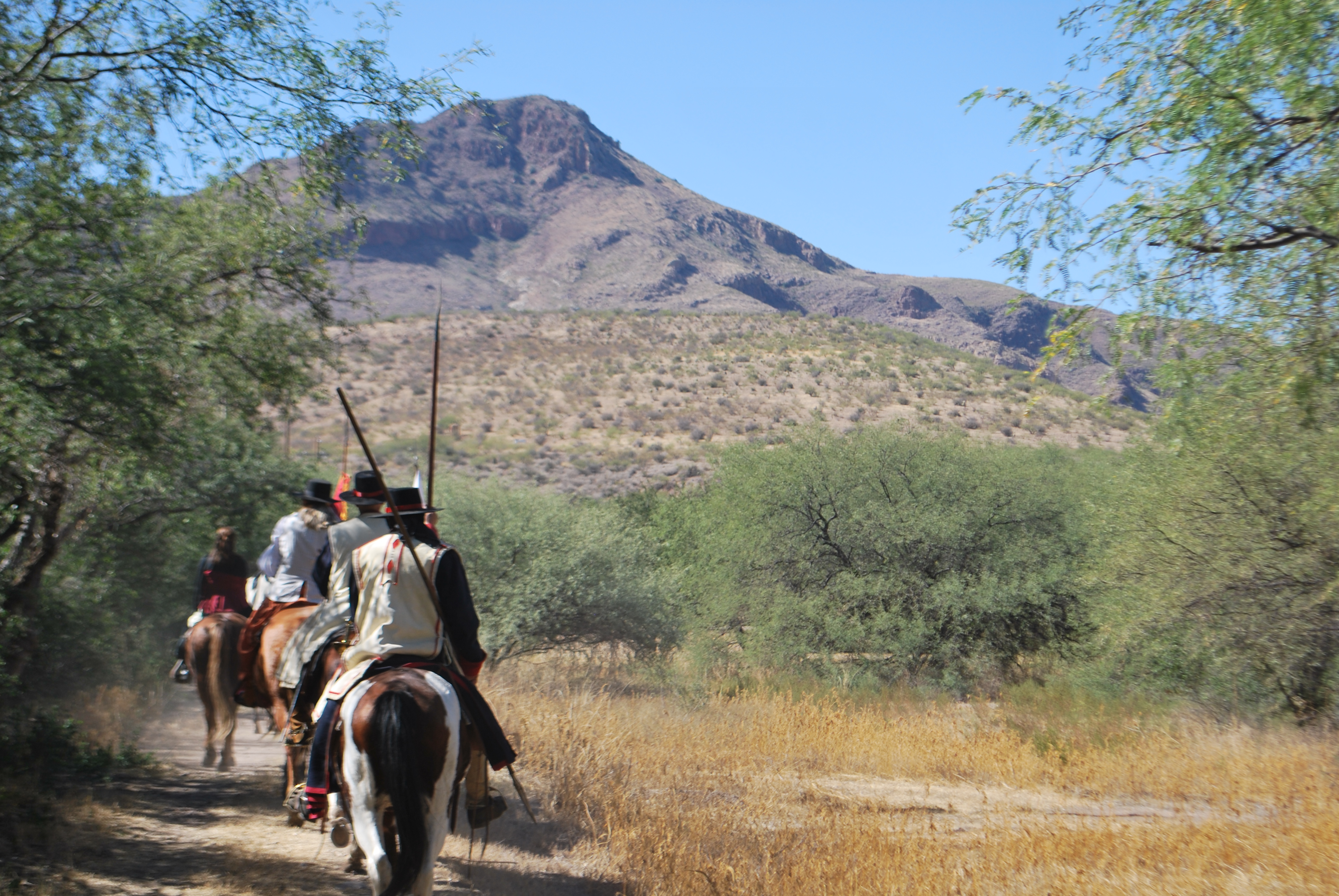 Living History - Mounted colonial Spanish soldiers ride the Anza Trail in a riparian desert landscape towards a mountain.