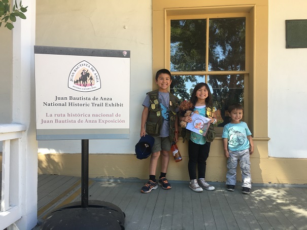 Three children standing in front of a window with Junior Ranger Badges and vests. 