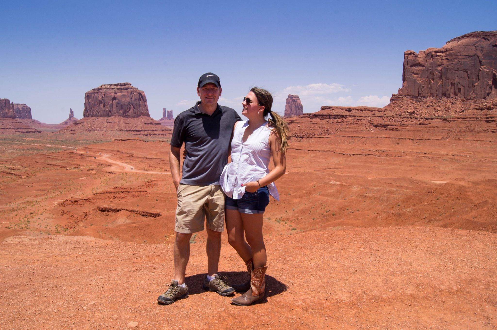 A man and a woman stand in front of the characteristically red-orange landscape of Monument Valley. In the background are buttes, rocky pillars, and cliffs.