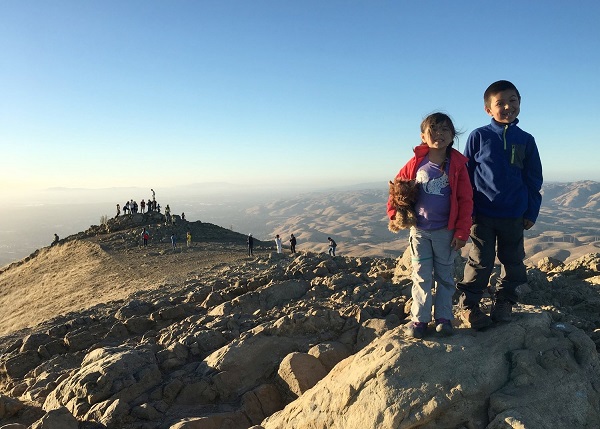 Two kids stand at a rocky peak above hills in the distance.