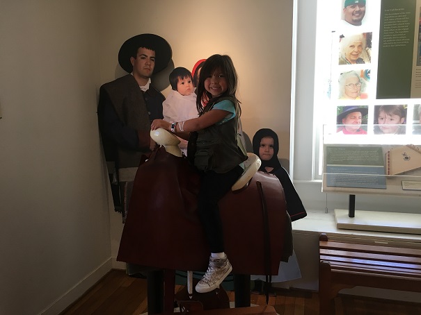 Young girl sitting on a wooden horse and saddle by an exhibit. 