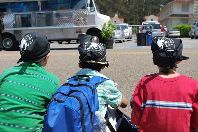 Three kids wearing Anza Trail bandanas on their heads rest on a street curb in front of a food truck