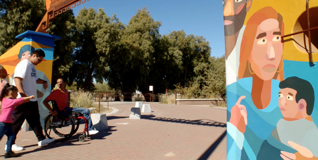 A man in a wheelchair and his friends enter a park through a colorful archway with a mural of a family