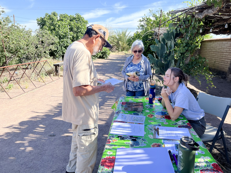 A young woman sits at a table outside in a desert garden and speaks to two adults standing in front of the table