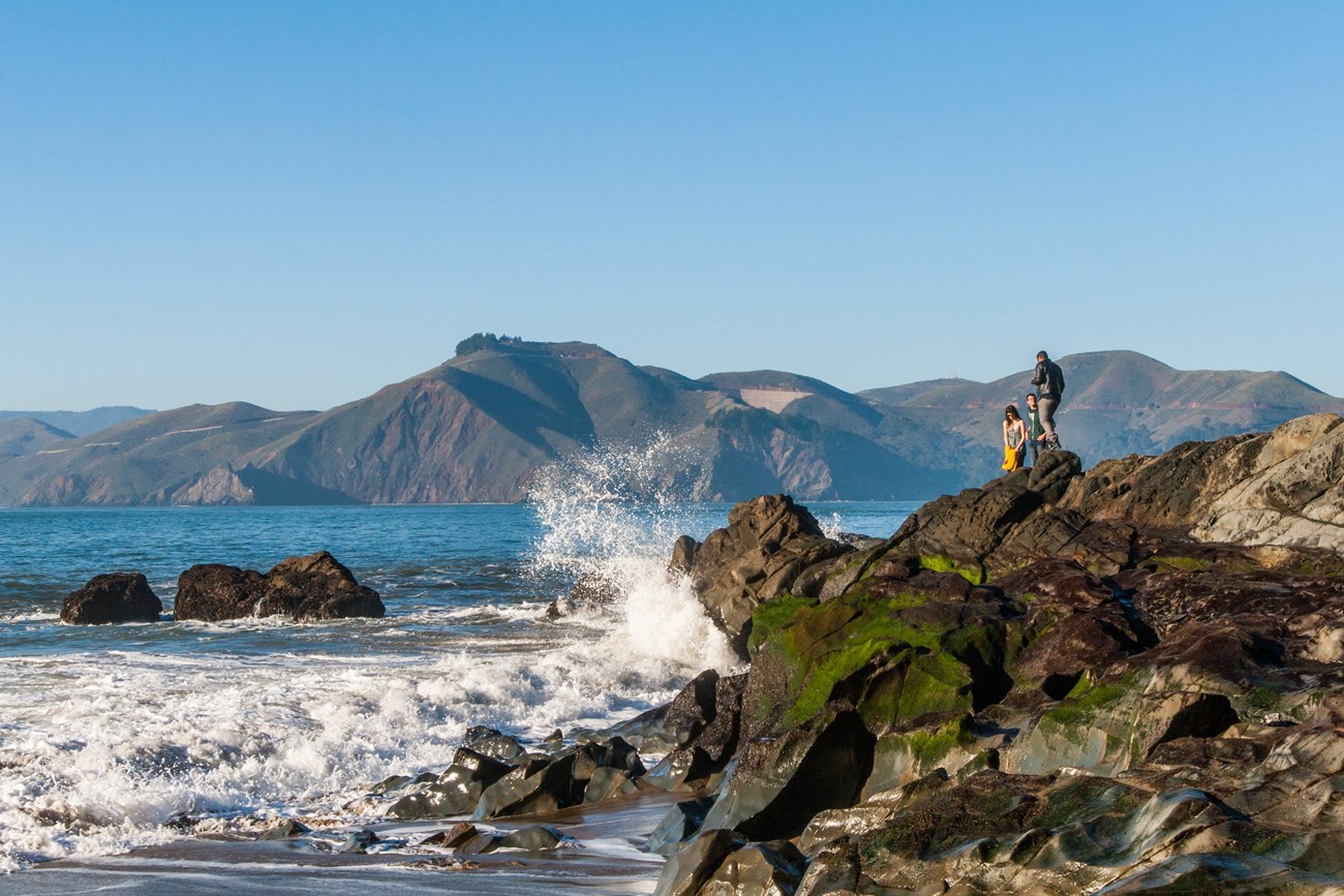 Waves crash against large jagged rocks in the San Francisco Bay with green round Marin Headlands in the distance