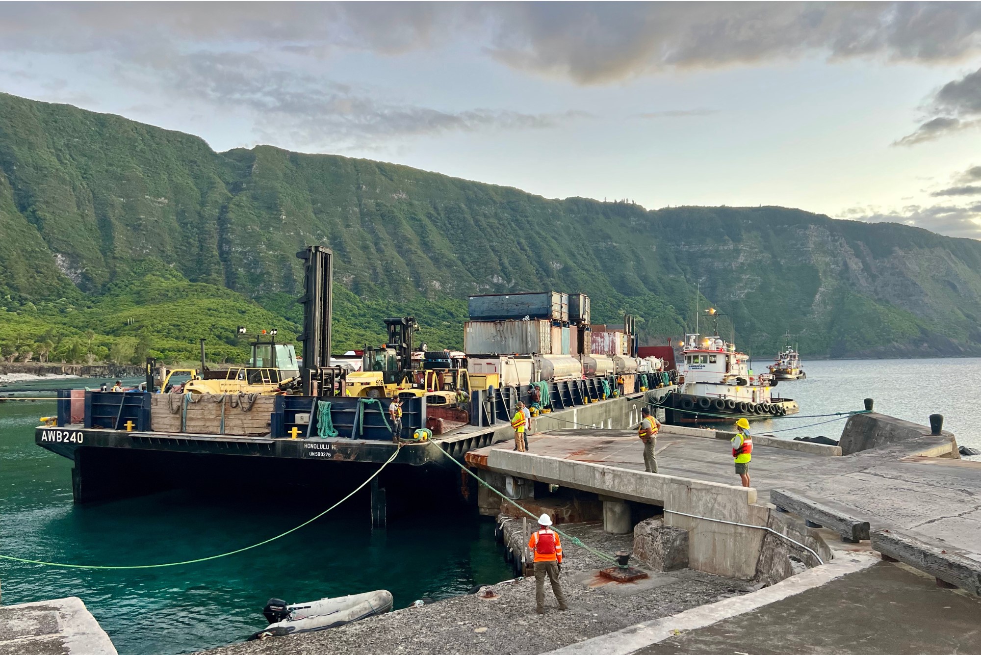 Outdoors; loaded barge at cement dock with workers and green cliffs behind.