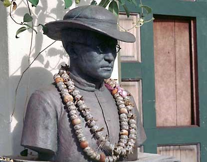 A metal statue of a man depicting Father Damien. He wears a hat and a few leis.