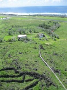 A view overlooking a road, a few houses, vegetation, rock walls, and an ocean in the distance.