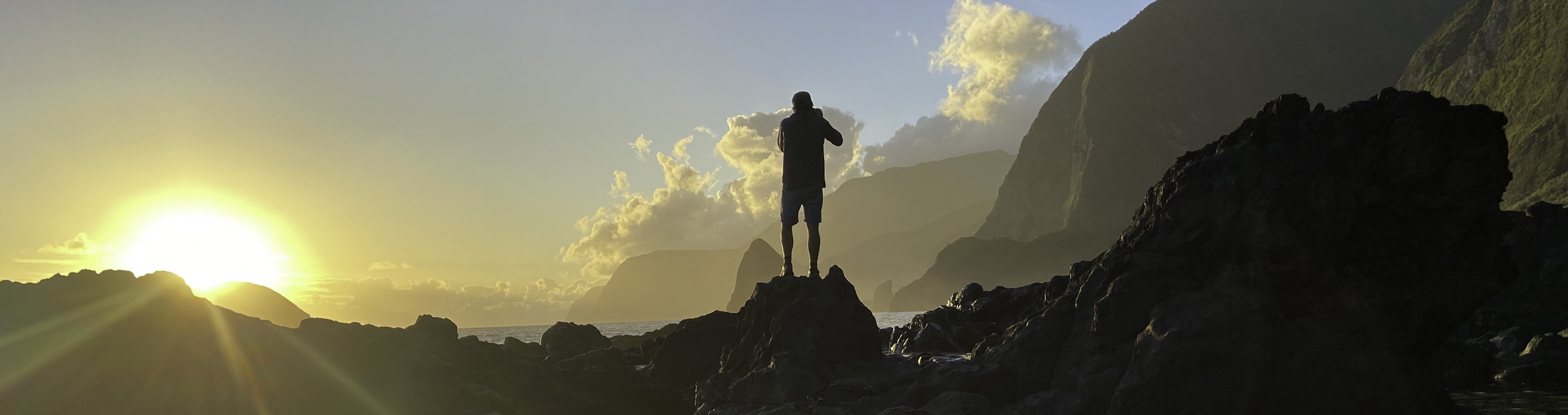 Outdoors; silohuette of person standing on rock taking photo, sun shining on left.
