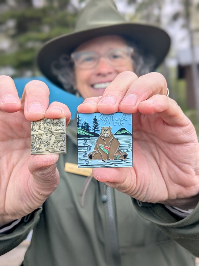 A photo of a ranger holding two pins, one is bronze the other is colored enamal
