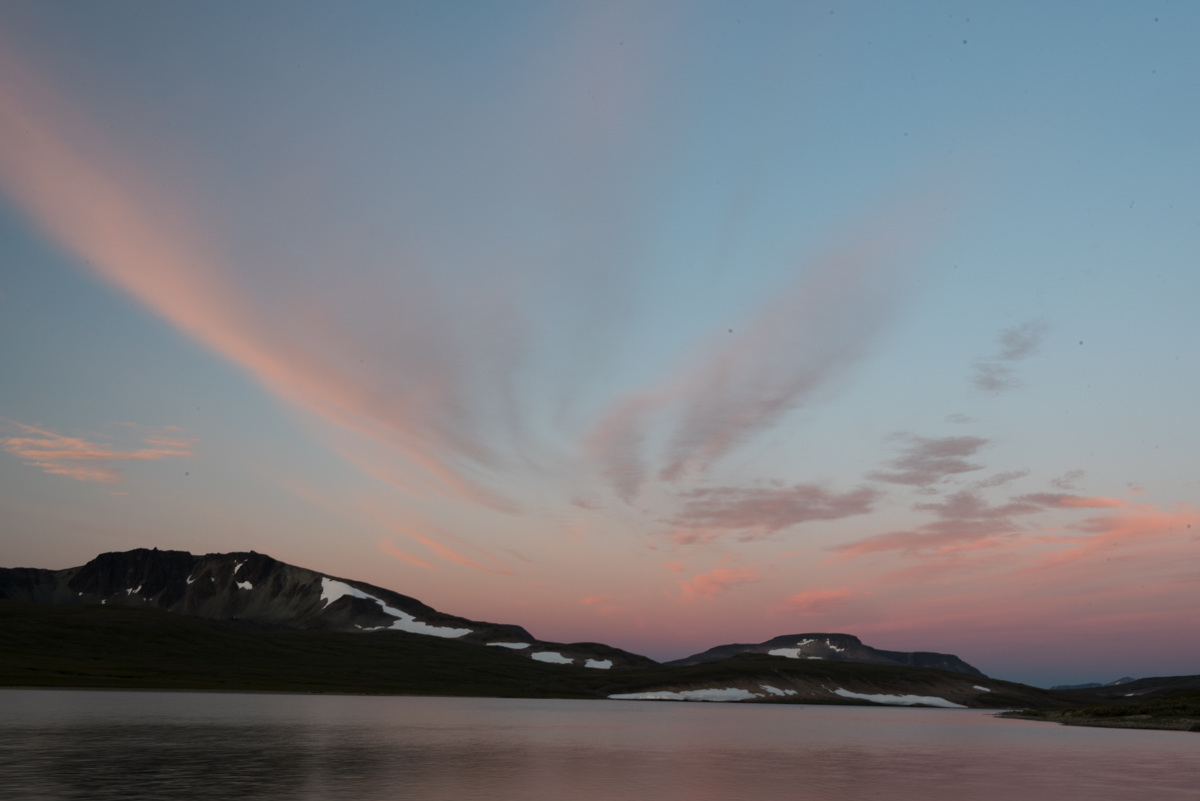A lake with snow patches on the mountains and pink clouds in the sky