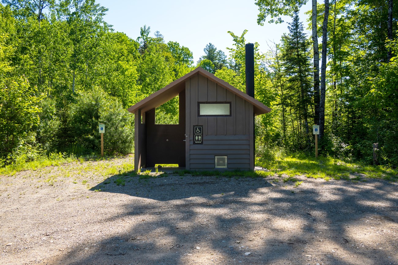 Two ADA parking spaces next to a vault toilet structure near a gravel road. A mixed forest is behind the parking spaces and vault toilet.