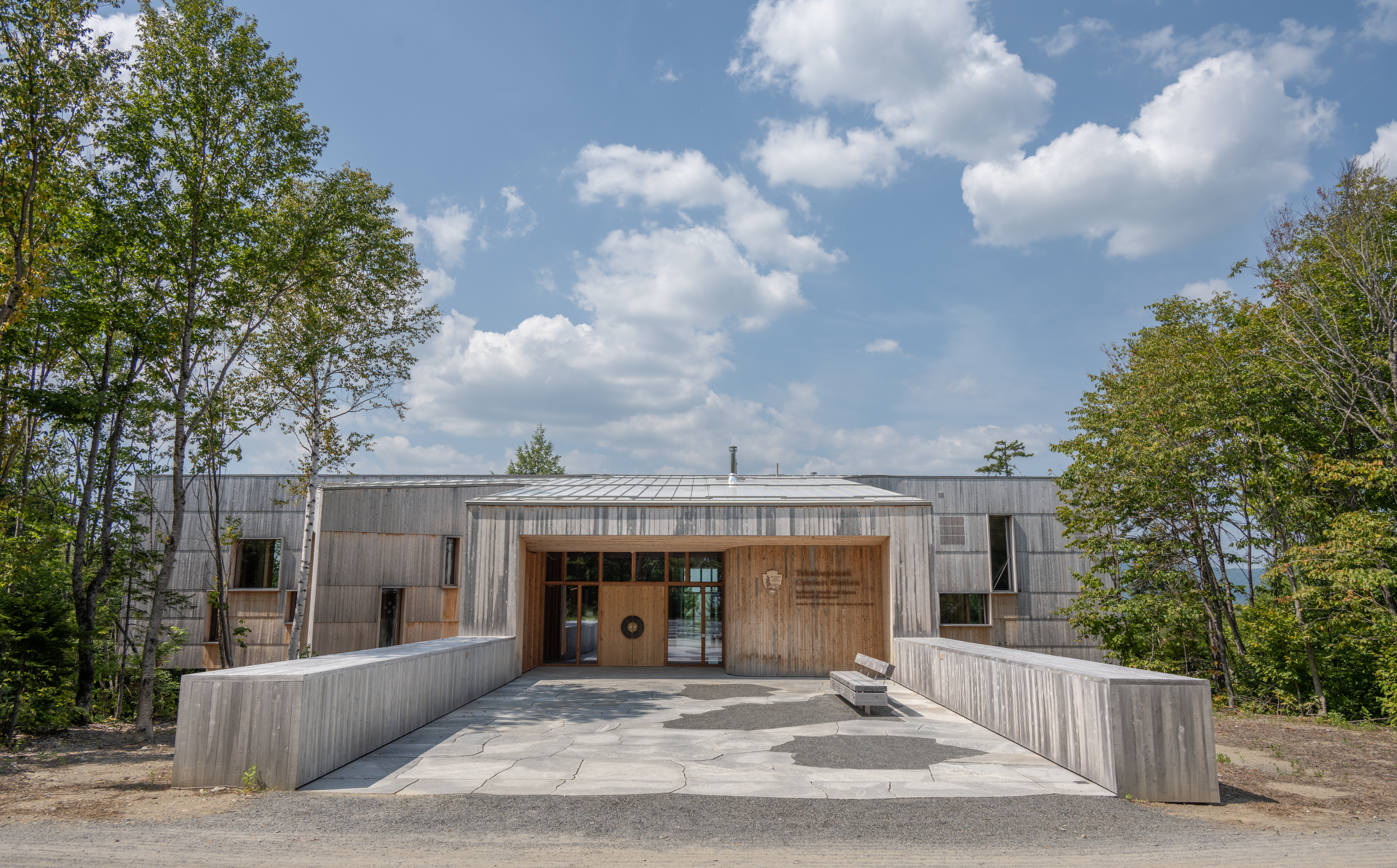 A large wooden bulding sits under a blue sky with puffy clouds and green trees on either side.