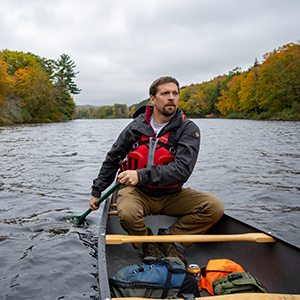 Man canoes down a dark river with fall foliage lining the shore.