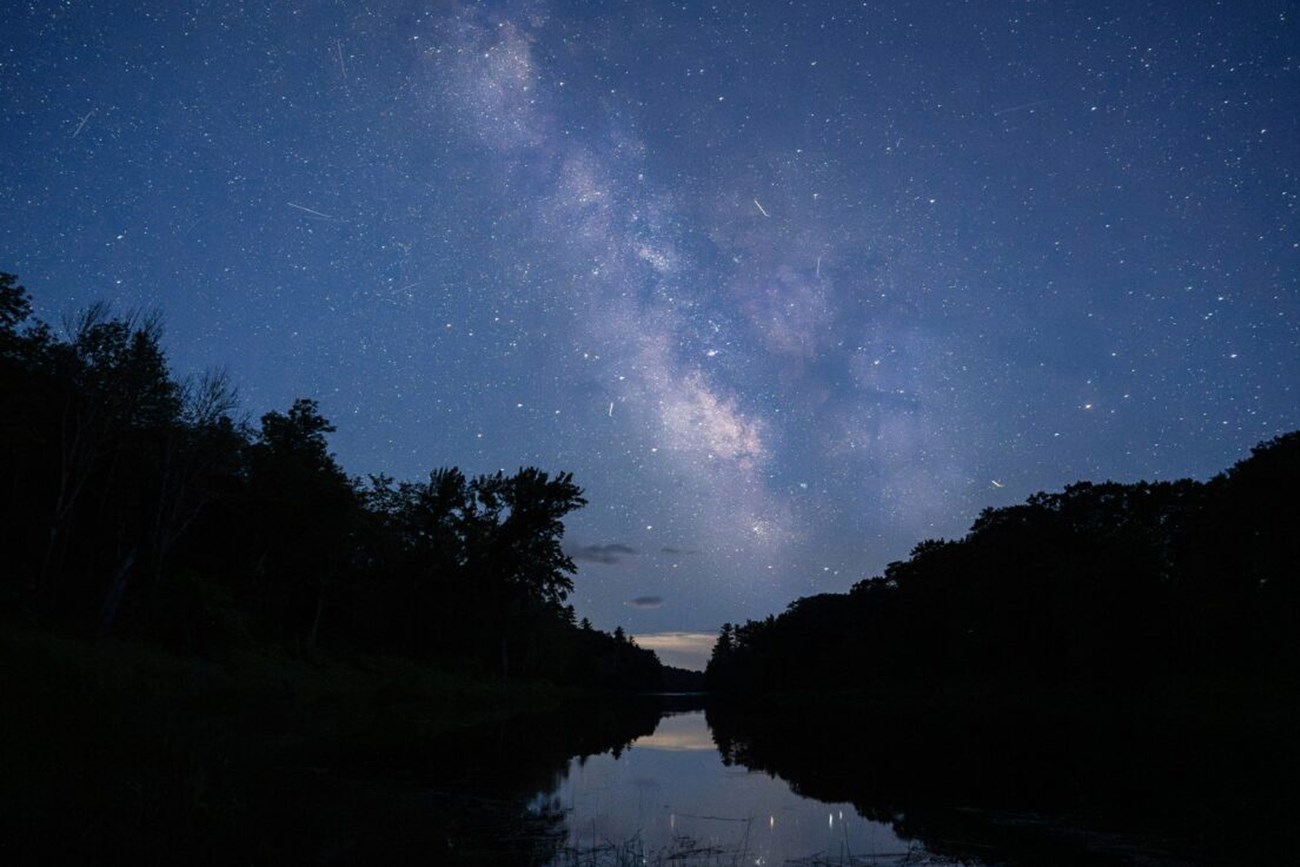 The Milky Way illuminated clearly over the East Branch Penobscot River. Silhouettes of the woods and landscape line both sides of the river. The Milky Way and the bright stars reflect onto the serene river.