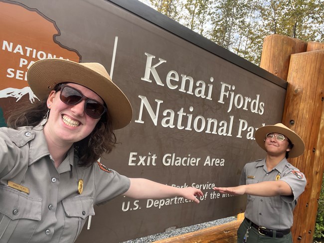 Two park rangers in uniform reach out an arm towards each other in front a park sign reading "Kenai Fjords National Park / Exit Glacier Area"