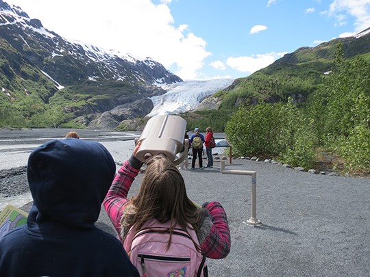 Children look through large binoculars at a glacier in the distance.