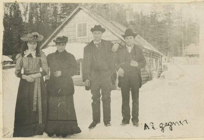 A historical black and white photo of four people standing in front of a cabin