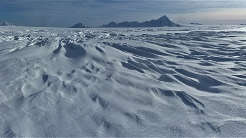 a large area of ice that is covered by snow.  Mountain peaks appear in the distance.