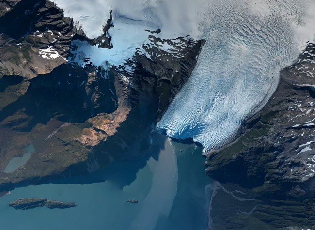 An aerial view of  Aialik Glacier.  The glacier starts from the Harding Icefield in the top right of the picture, and flows to the middle.  The glacier is surround by land on the right and left, and water at the bottom of the image.