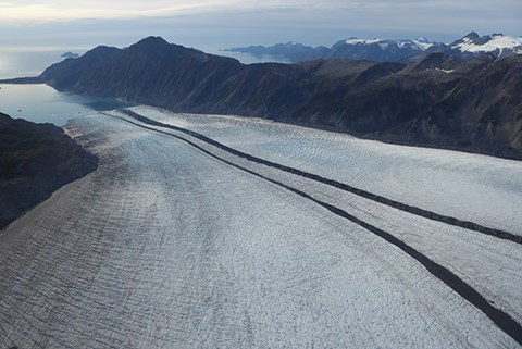 A large white glacier with two dark lines running down the center.  In the upper left of the image the glacier meets the ocean, and in the background are mountains