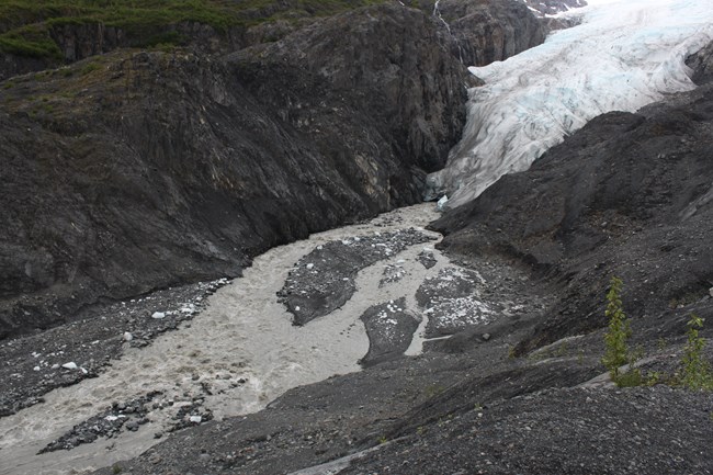 A glacier flows from the upper right of the image towards the bottom left.  The glacier ends in the middle of the image, and a river of meltwater continues to the bottom left.  Above and below the glacier is a rocky mountainside