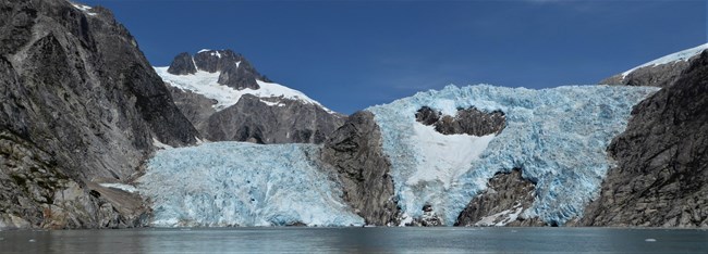 A blue glacier is in the valley between two mountainsides.  There are patchs of rock showing underneath the glacier.  In the background is a mountain with snow near the top.