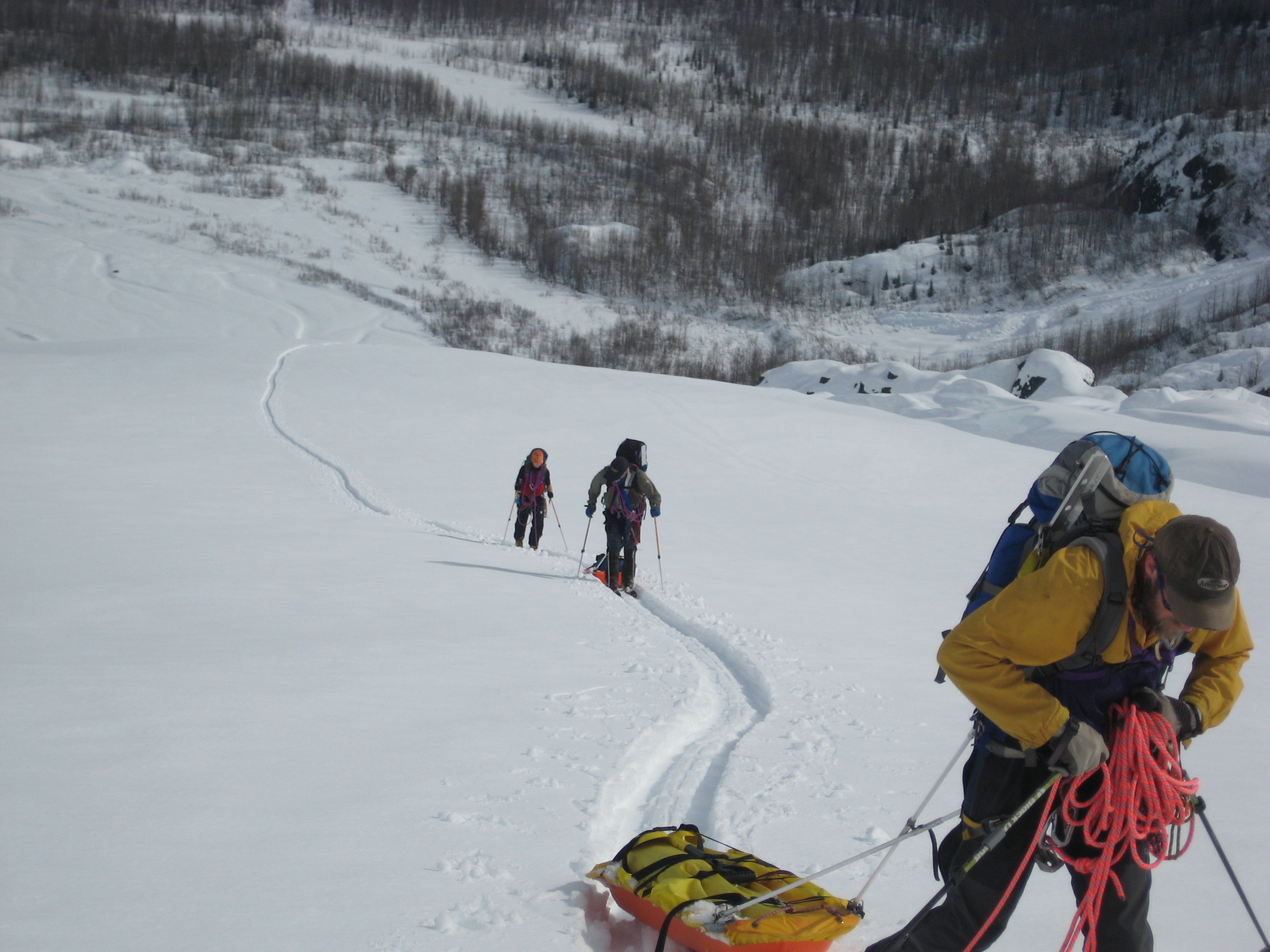 Three people with safety gear ski up a snowy mountainside
