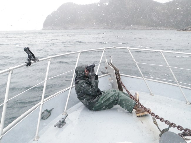 A person in rain gear looks through binoculars while on a boat bow in the rain.