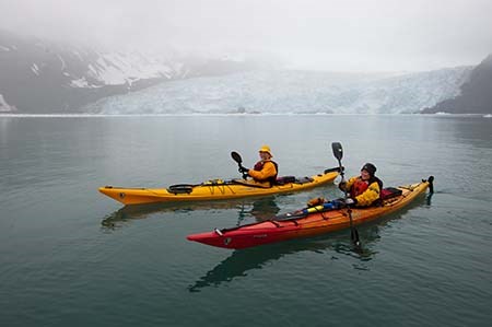 Two kayakers sit in the water in front of a large tidewater glacier.