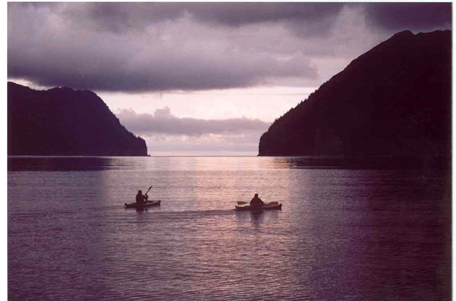 Two kayakers float in the ocean, framed by steep shorelines.