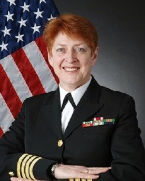 Portrait of female with short red hair in uniform in front of American flag. Wears a formal black jacket with white blouse underneath. Jacket has 3 yellow stripes at end of long-sleeves and several colored bars pinned on jacket chest.
