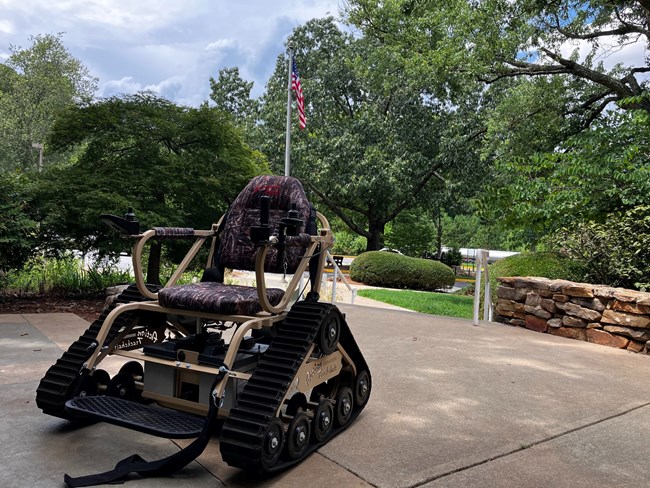 All terrain wheelchair sits on patio with trees and American flag behind.