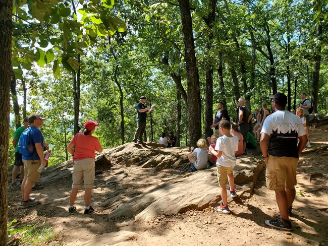 Group of people gather outside listening to a ranger talking. Ranger stands on top of rock. Trees surround.