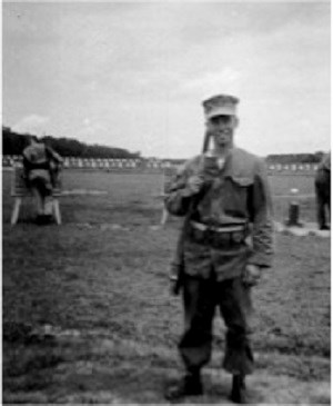 White-skinned male stands outside in uniform surrounded by flat land with sporadic dried grass. He wears baggy pants tucked into boots. Long-sleeve, button down shirt with ammunition belt over waist.