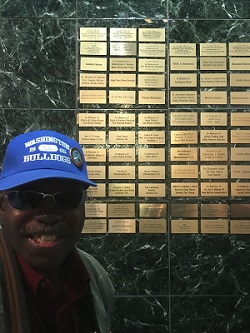 Black man in blue baseball cap and glasses smiles next to a plaque of gold-plated names on a wall.