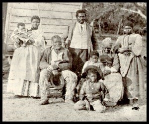 Black and White photo: 2 adult males, 3 adult females, 2 young boys, and a baby pose in front of a house. All are black individuals.