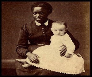 black woman in a collared dress sits with a white toddler in a dress on her lap.