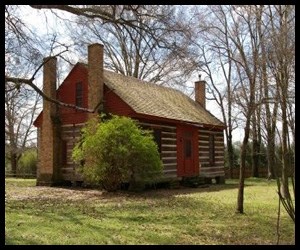 1 story log cabin with 2 chimneys on 1 side and 1 chimney on the other and a triangle roof. Scattered dormant trees surround the house outside.