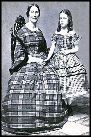 A young girl stands next to a seated adult female. They both wear dresses with large petticoats.