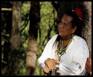 Man with dark skin and hair pulled back leans against a tree. He wears several beaded necklaces and a small spiky headress on his head