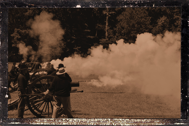 Sepia-toned image of cannon crew firing a cannon. Smoke shoots out of cannon from left to right. People in Civil War uniforms stand around cannon at left.