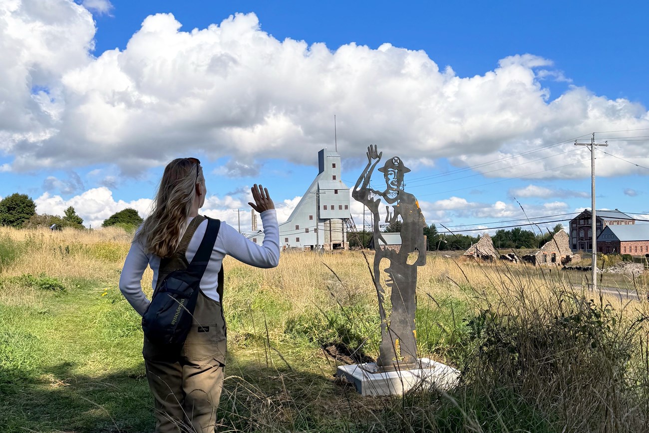 A woman faces away from the viewer looking towards a metal sculpture with tall mining buildings in the distance.