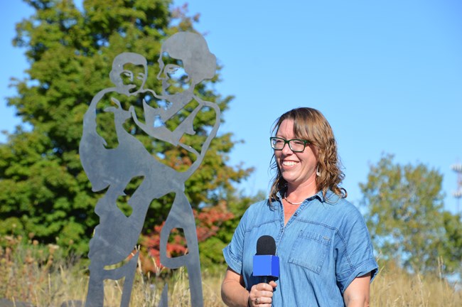 Artist Kasey Koski holds a microphone and smiles standing next to the woman and baby sculpture