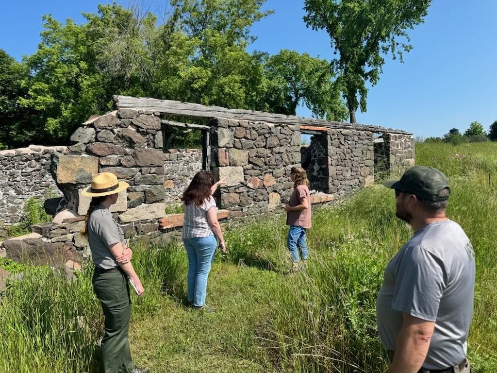 Multiple people stand in tall grass facing away from the viewer looking at the foundation of a ruined building.