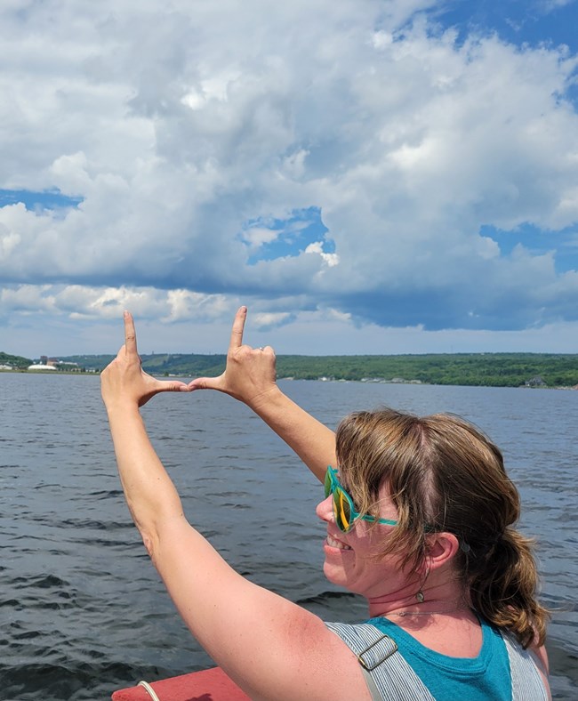 A woman wearing sunglasses looks off to the side while holding her hands up with a large lake in the background.